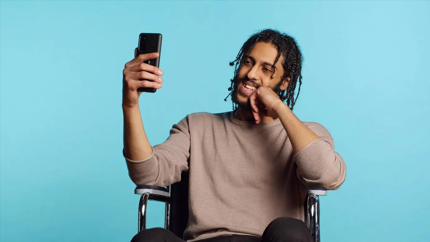 Hombre en silla de ruedas saludando a un amigo por una videollamada en línea en un teléfono móvil, en estudio.